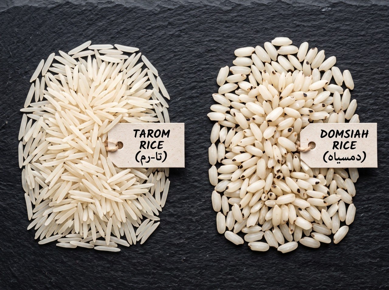 Two piles of Iranian long-grain rice side by side on dark slate, one labeled tag Tarom and one Domsiah with dark-tipped grains, top-down macro shot, natural soft light