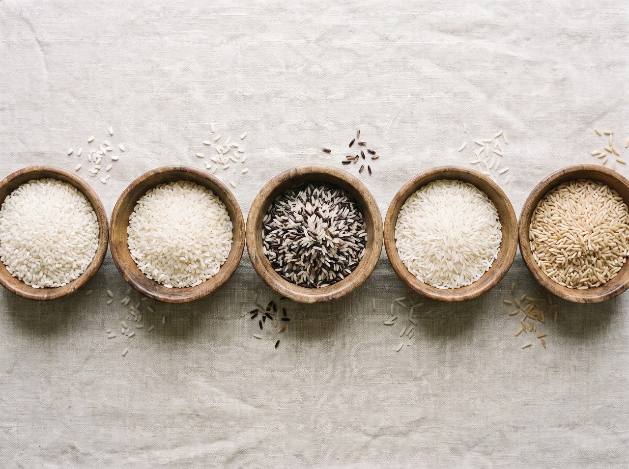 Comparison of five Iranian northern rice varieties in separate wooden bowls on a linen surface, top-down studio shot, natural light